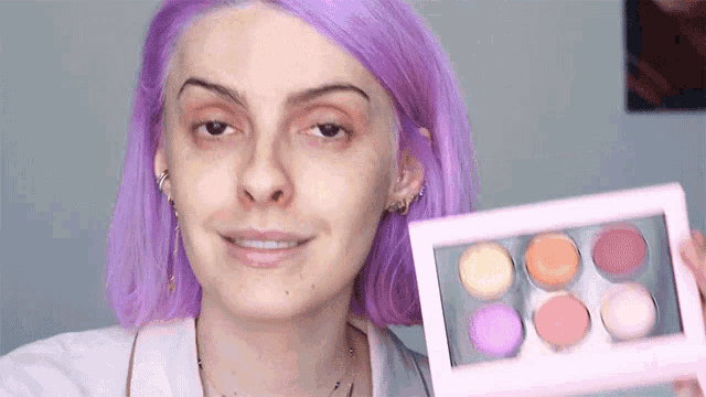 a woman with purple hair is holding a pink palette of makeup .