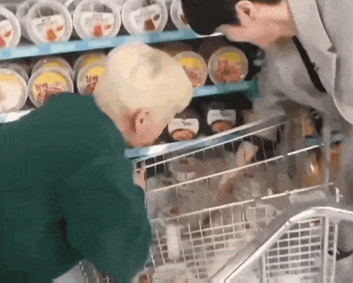 two men are shopping in a grocery store and one of them is looking at a cart filled with food .