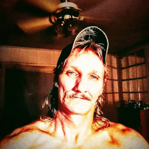 a shirtless man with long hair and a hat looks at the camera in front of a ceiling fan