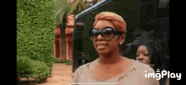 a woman wearing sunglasses is standing in front of a bus with imgplay at the bottom of the screen