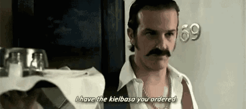 a man with a mustache and a white shirt is standing in front of a table .