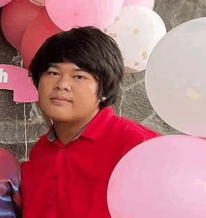 a man in a red shirt is standing in front of a wall surrounded by pink balloons .