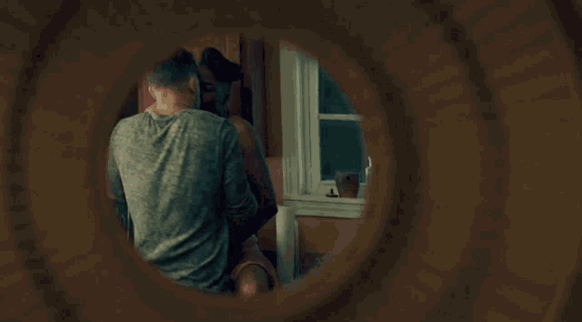 a man in a grey shirt is looking out a window through a hole in the wall