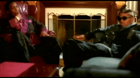 a man in sunglasses sits next to another man in a chair in front of a fireplace
