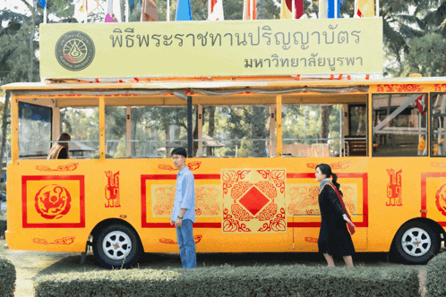 a man and woman are standing in front of a yellow bus with a sign above it that says ' มหาวิทยาลัย บูรพา '