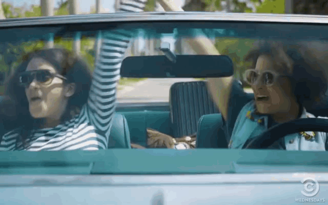 two women are driving a car with their arms in the air and the rear view mirror says wednesdays