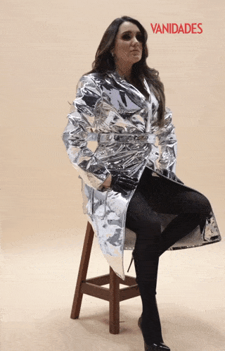 a woman is sitting on a wooden stool wearing a silver coat with vanidades written on it