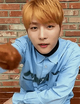a young man with blonde hair is wearing a blue shirt and holding a chicken wing in front of a brick wall .