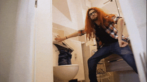 a man with long red hair and glasses is standing in a bathroom next to a toilet