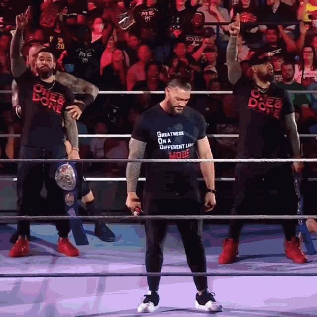 a man in a black shirt that says " greatness on a different level " stands in a wrestling ring