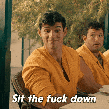 a man in a yellow shirt sits at a table with the words sit the fuck down above him
