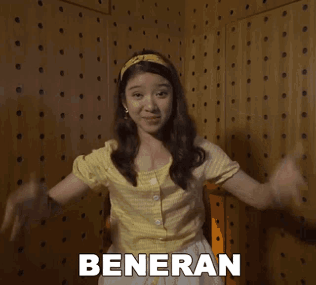 a woman in a yellow shirt and white skirt is standing in front of a wooden wall with the word beneran on it