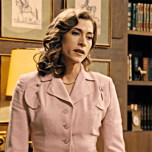 a woman in a pink jacket and pearl necklace stands in front of a bookshelf