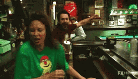 a woman in a green shirt is dancing in a bar while a man in a red shirt is standing behind her .