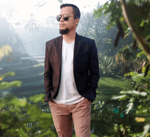 a man wearing sunglasses and a suit jacket stands in front of a lush green field