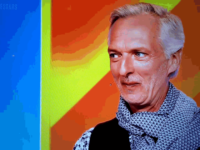 a man with gray hair and a scarf looks at the camera with the words estars in the corner