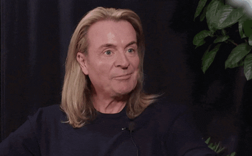 a man with long blonde hair is smiling with a dark background