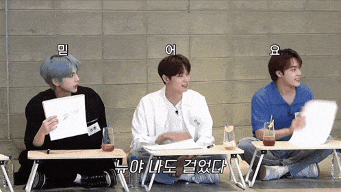 a group of young men are sitting at small desks and one of them is holding a piece of paper that says " i love you "