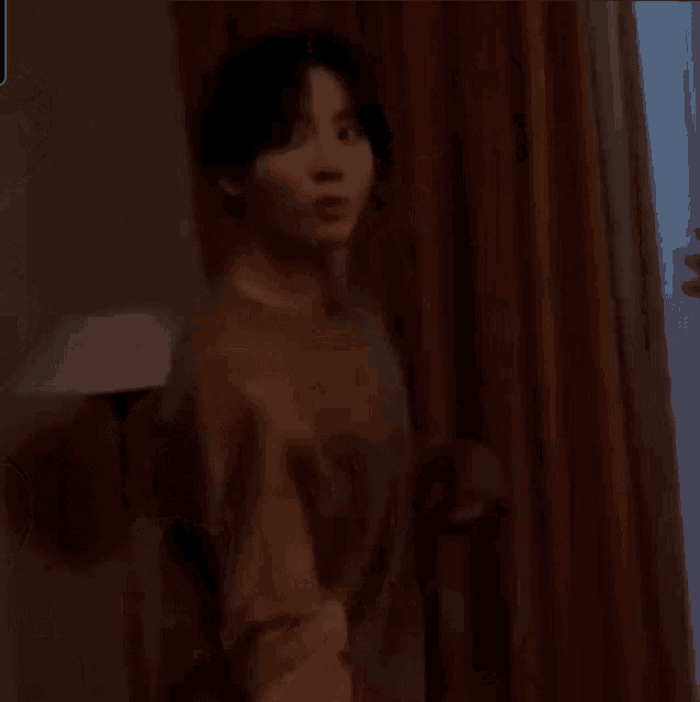 a man in a brown shirt is standing in a dark room next to a window .