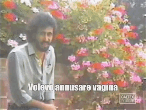 a man with a beard is sitting in front of a bush of flowers and says volevo anusare vagina