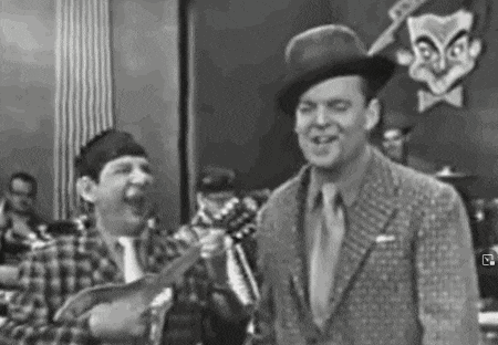 a man in a suit and tie is holding a guitar and laughing while another man holds a guitar .