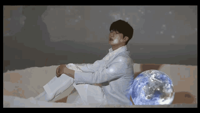 a man in a white suit sits next to a globe with his eyes closed