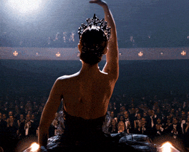 a woman in a black dress with a crown on her head is dancing in front of a crowd