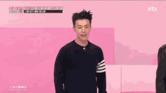 a man in a black sweater stands in front of a pink wall with a tbs logo on it