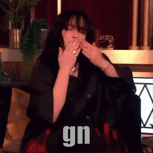 a woman is sitting on a couch and covering her mouth with her hands and the word gn is on the bottom right