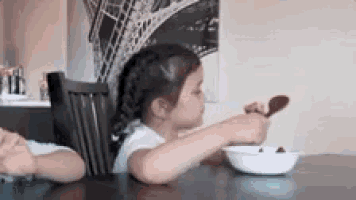 a little girl is sitting at a table with a bowl of cereal and a spoon .