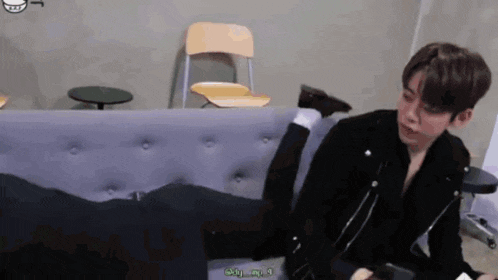 a young man in a black jacket is sitting on a couch with his legs crossed .