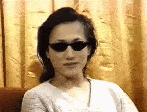 a woman wearing sunglasses and a white sweater is sitting in front of a yellow curtain .