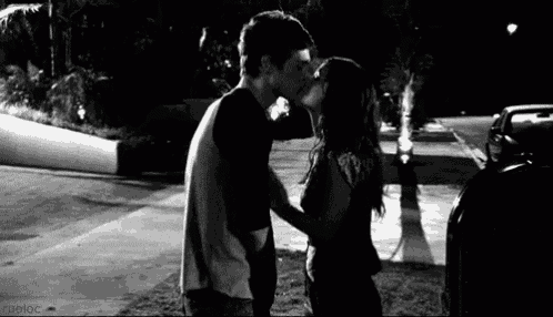 a black and white photo of a man and a woman kissing on the side of the road .
