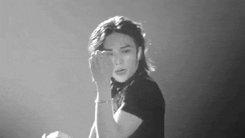 a black and white photo of a man with long hair covering his face with his hand .