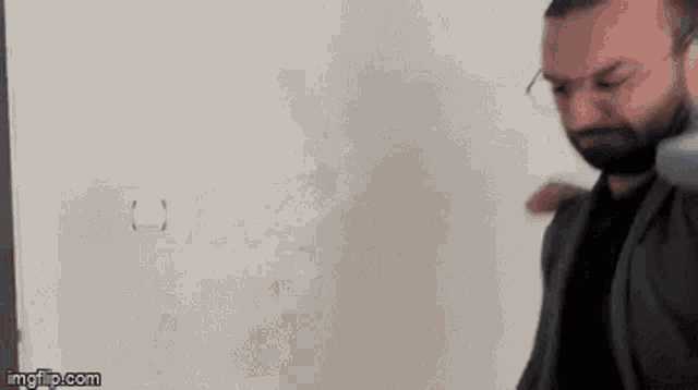 a man with glasses is standing in front of a white wall .