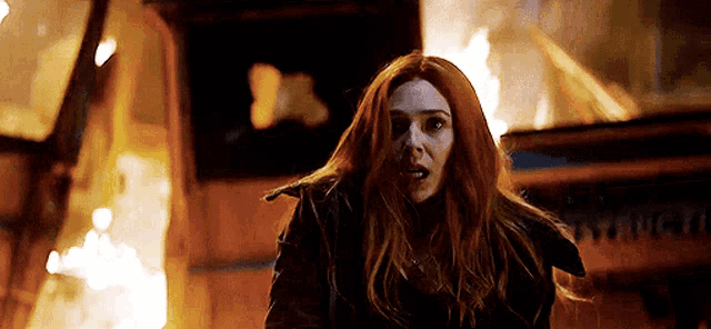 a woman with red hair is standing in front of a building that is on fire .