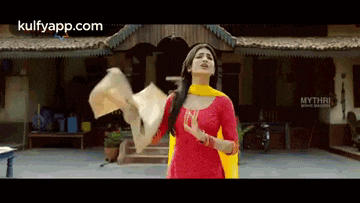 a woman in a red dress and yellow dupatta is standing in front of a building holding a piece of paper .