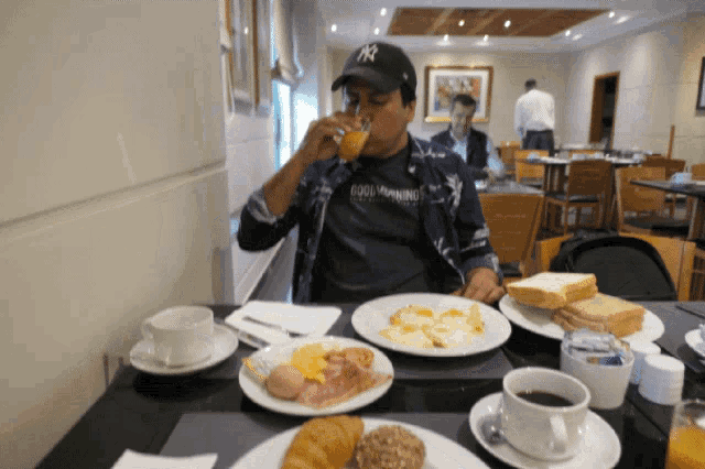 a man wearing a ny hat sits at a table with plates of food