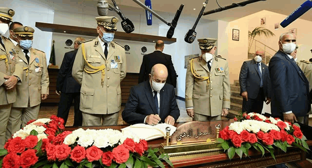 a man wearing a mask is signing a book with flowers in front of him