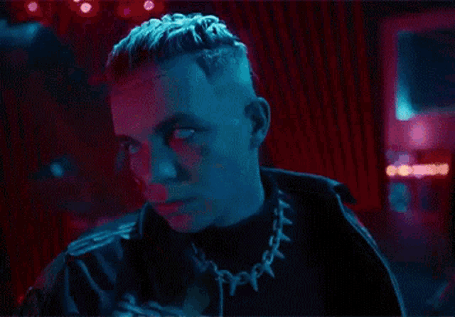 a young man with a tattoo on his face is wearing a chain around his neck and looking at the camera .