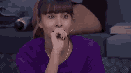 a woman in a purple shirt is sitting on a couch covering her face with her hand .