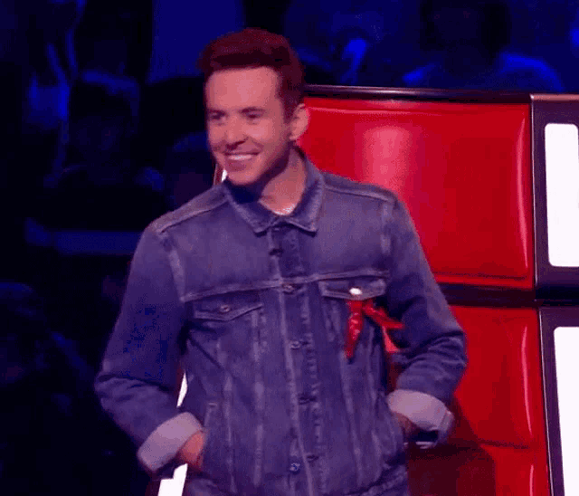 a man wearing a denim jacket is standing in front of a red chair .