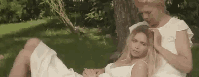 two women in white dresses are sitting next to each other in the grass under a tree .
