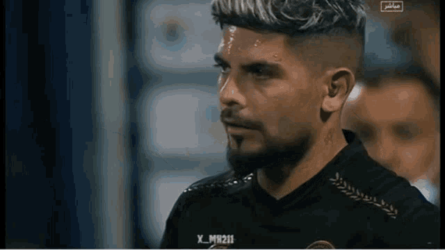 a man with a beard and a tattoo on his face is wearing a black shirt with arabic writing on it .