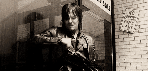 a man in a leather jacket is holding a gun in front of a building with a no parking fire lane sign .