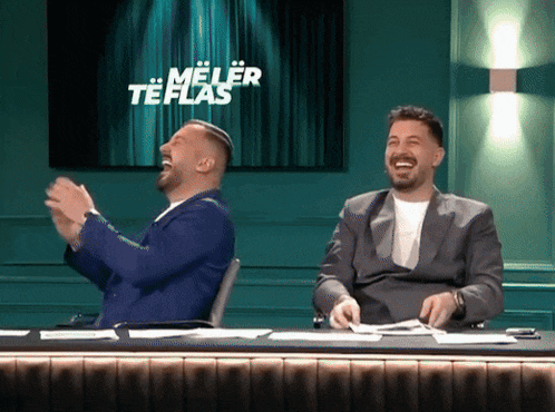 two men are sitting at a table laughing in front of a screen that says meler te flas