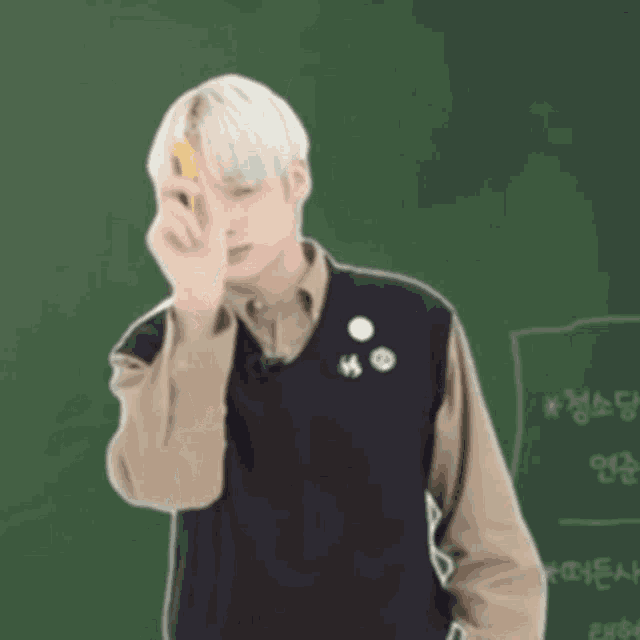 a young man is standing in front of a green chalkboard holding a yellow eraser to his eye .
