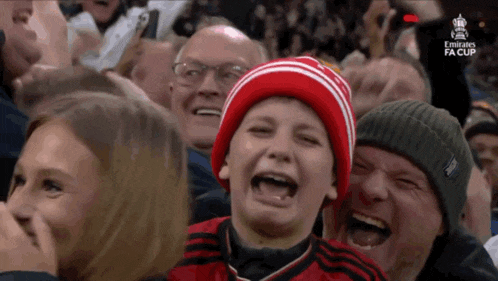a boy in a red hat is laughing in a crowd with the emirates fa cup written on the screen