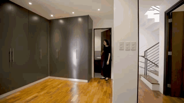 a woman in a black dress stands in an empty room with a staircase behind her