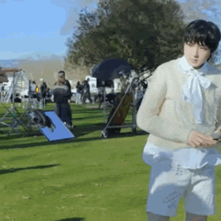 a woman in a white sweater and white shorts is standing in a grassy field holding a cell phone .